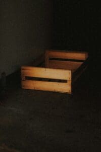 A wooden bed in the dark with no sheets.